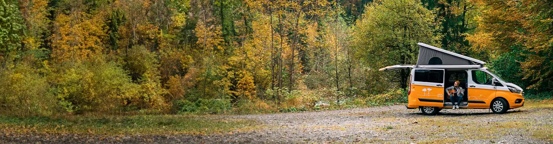 Man with guitar sitting in the doorway of an orange campervan parked on a graveld surface in the forest at autumn