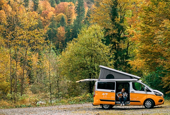 Man with guitar sitting in the doorway of an orange campervan parked on a graveld surface in the forest at autumn