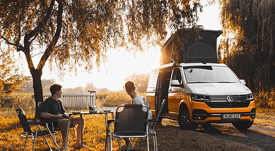 Two men with their backs turned towards the camera, sitting on camping chairs under a tree next to a yellow and white van during sunset