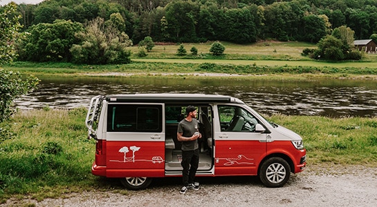 Man standing in front of a red campervan parked right next to a river.