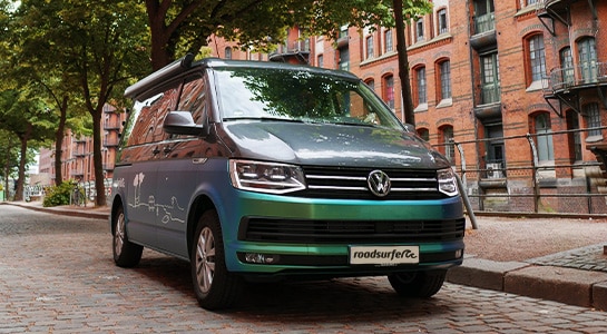 Metallic green campervan in front of a brick house parked on the street.