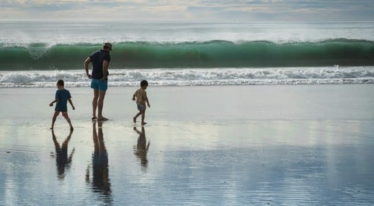 Adult with two kids walking along wet sand as a green wave curls in—things to do in California with kids.