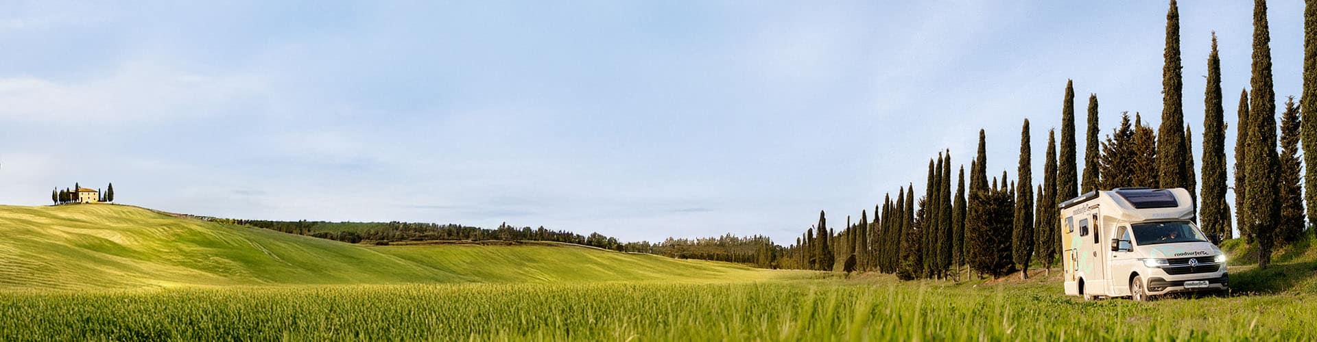 A motorhome is driving along a scenic countryside road lined with tall cypress trees, surrounded by green fields and clear skies