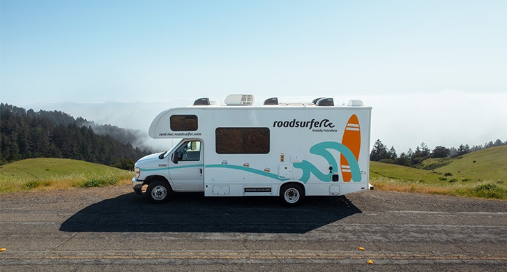 A motorhome is parked on a scenic mountain road with rolling green hills and mist in the background.