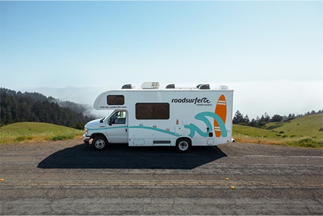 A motorhome is parked on a scenic mountain road with rolling green hills and mist in the background.