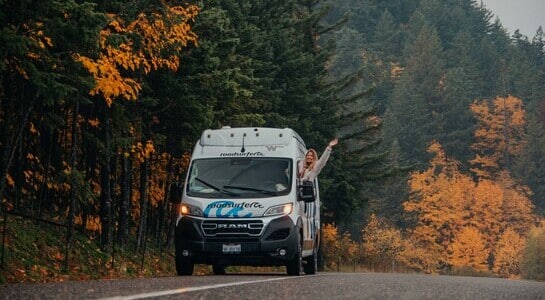 Woman leaning out of the window of her roadsurfer motorhome standing on a road with a autumn scenery