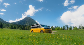 A new VW California camper driving on a road through green fields with mountains in the back