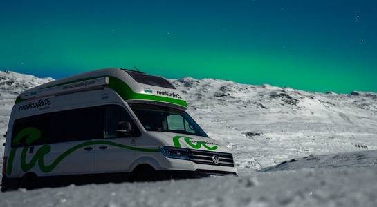 Green and white motorhome from roadsurfer standing in the snow with northern light in the sky