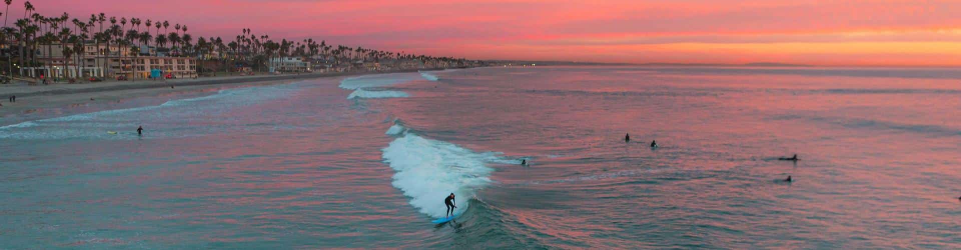 Sunset surf at Oceanside with surfers paddling out, pastel sky, and palm-lined coast—showcasing the beauty of surf beaches in California