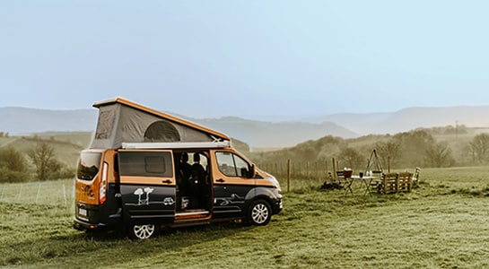 Orange and dark blue campervan with rooftop tent parked on a vineyard overlooking wide green fields and hills.