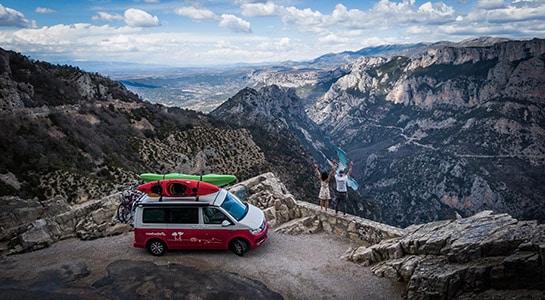 Mountain panorama, couple throwing their hands in the air next to a red and white van with kayaks on its roof