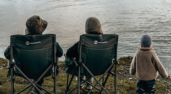 Man and woman sitting in camper chairs next to a child in front of a river.