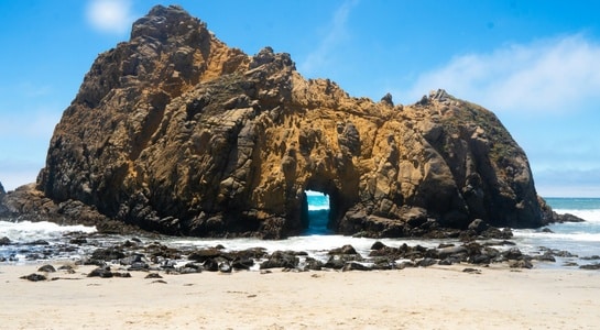 Sun shining through Keyhole Arch and unique rock formations at Pfeiffer Beach, a dramatic example of beaches in California’s Big Sur.