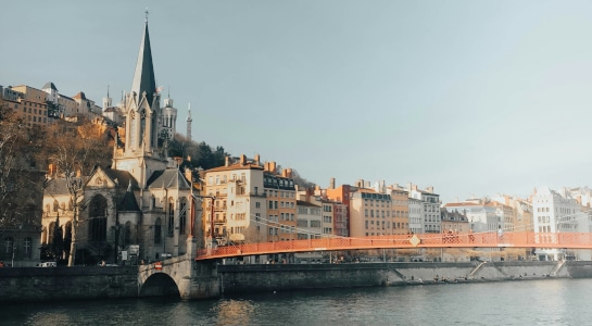 Picturesque riverside scene in Lyon, France, featuring the Saint-Georges Church, colorful buildings, and the iconic red Passerelle Saint-Georges footbridge bathed in soft sunlight