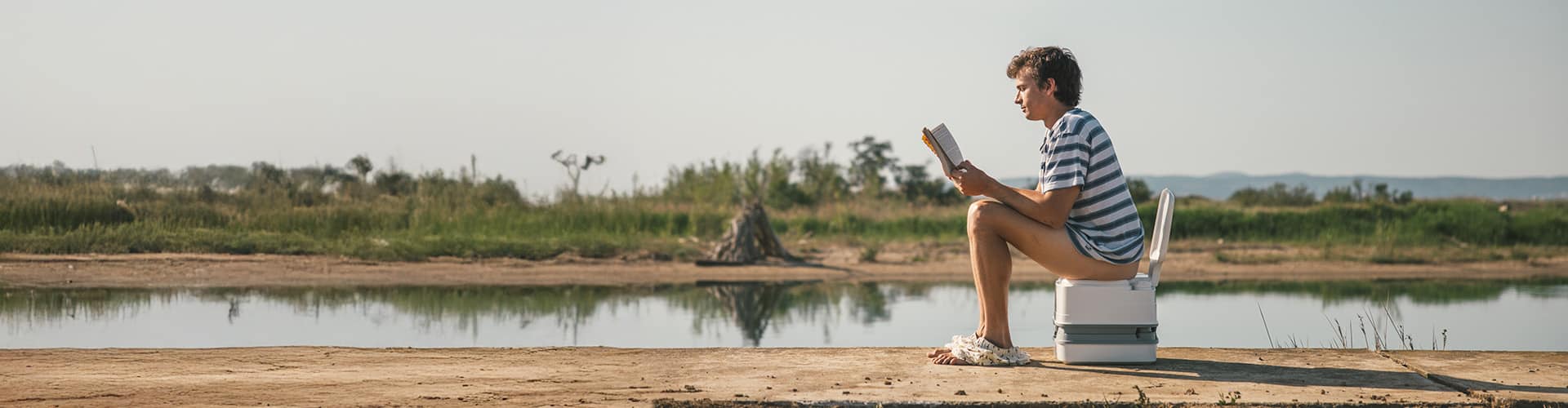 Person sitting on a portable camper toilet outdoors near a lake during a road trip.