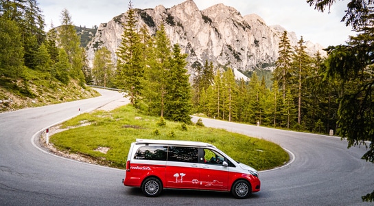 Red Mercedes Marco Polo camper takes a curve in a mountain road with mountain scenery in the background