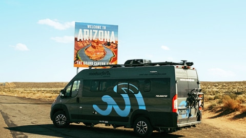 Grey roadsurfer boxvan photograph standing at the roadside in front of a Arizona state sign.