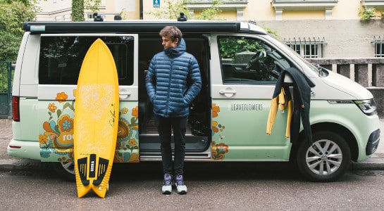 Young man with a yellow surfboard standing in front of a roadsurfer campervan