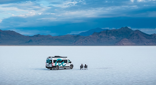 Two people sitting on camping chairs next to a white box van on the salt flats looking at the mountains.