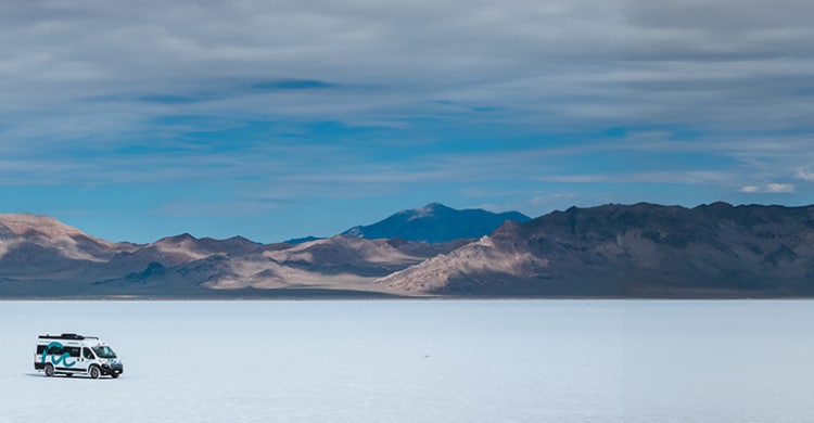 Box van driving on the salt flats in front of mountains during a cloudy day.