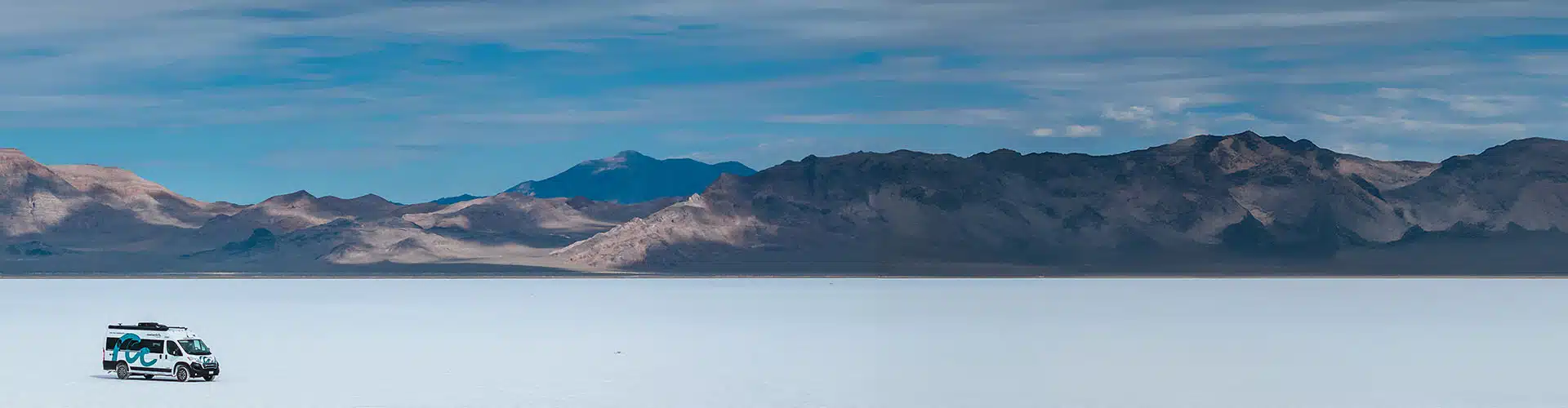 Box van driving on the salt flats in front of mountains during a cloudy day.