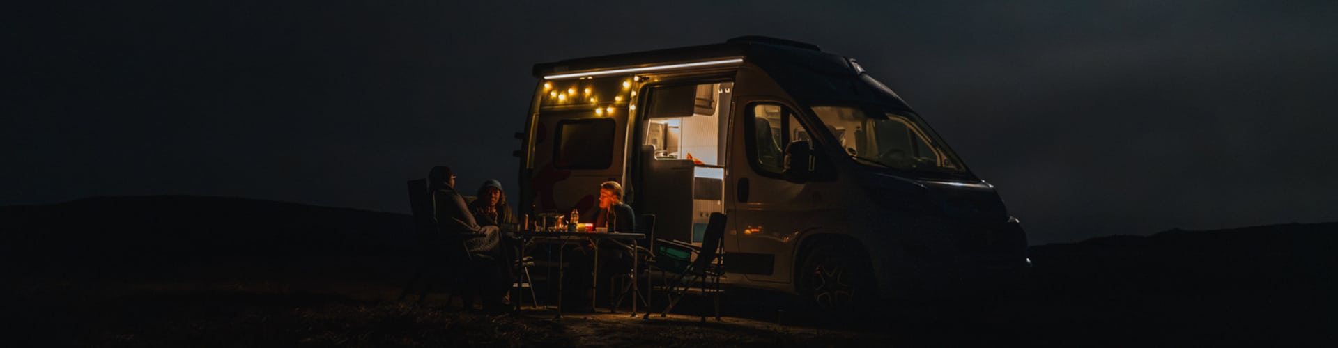 Three friends sharing dinner at a camping table next to a roadsurfer campervan at night, lit by warm string lights under a dark, misty sky.