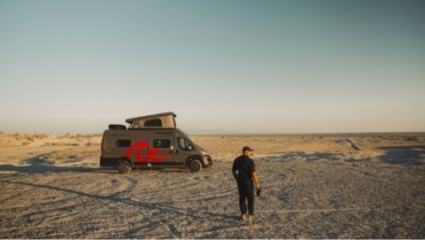 Man standing beside a roadsurfer campervan with its pop-top roof raised, parked on a pale desert flat