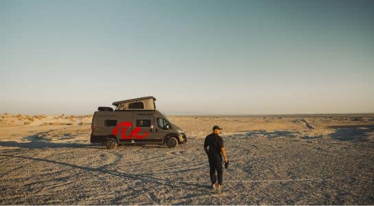 Man standing beside a roadsurfer campervan with its pop-top roof raised, parked on a pale desert flat