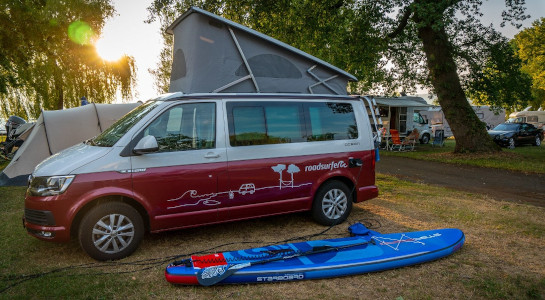 roadsurfer van camping with a blue SUP lying next to it