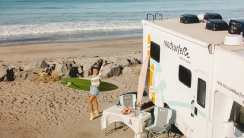 roadsurfer RV parked by the ocean with a surfer holding a board on a sunny Southern California beach