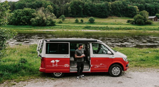 Man with a cup of coffee standing next to a red campervan, a river in the background
