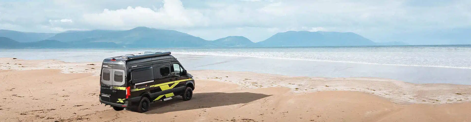 Black offroad Campervan standing on the beach at the sea with hills and clouds in the background