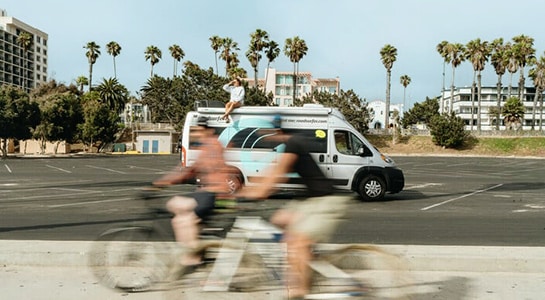 roadsurfer sprinter RV parked by the beach in California with palm trees in the background and people riding their bikes in the foreground