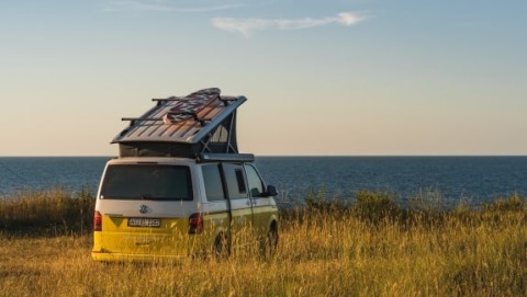 Yellow roadsurfer camper van with surfboards on the roof parked on a grassy clifftop above the sea
