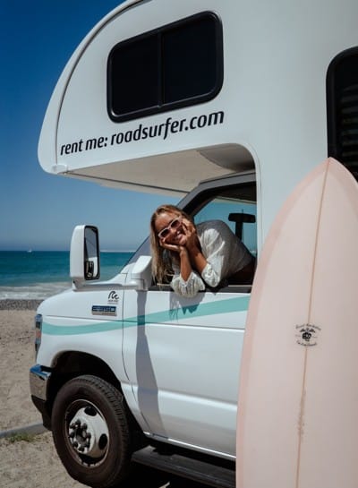 Woman leaning out of a Class C RV parked on a beach, with a surfboard in the foreground