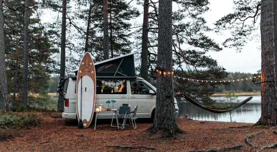 White van standing in a forest by a lake in the evening, surfboard leaning against the car, fairy lights and a hammock hanging between the trees
