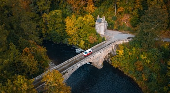 Camper van crossing an old stone bridge surrounded by autumn forest on a Scotland road trip in Royal Deeside