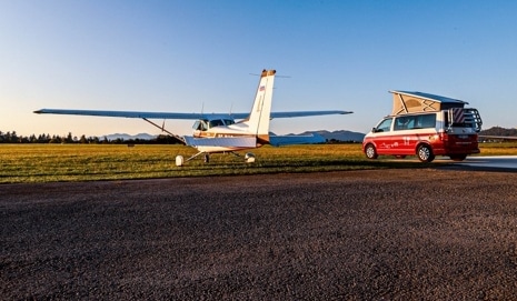 A red camper van with its roof raised parked beside a small airplane on a sunny airfield, both centered in the frame.