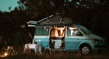 RV camping scene with a camper van set up for the evening, outdoor table and chairs, and lights at a campsite