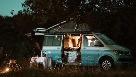 RV camping scene with a camper van set up for the evening, outdoor table and chairs, and lights at a campsite