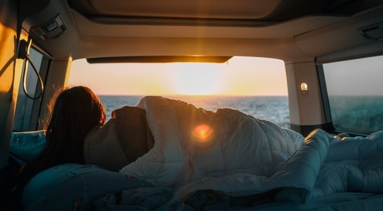 RV camping scene with a person resting inside a camper van at sunrise by the ocean