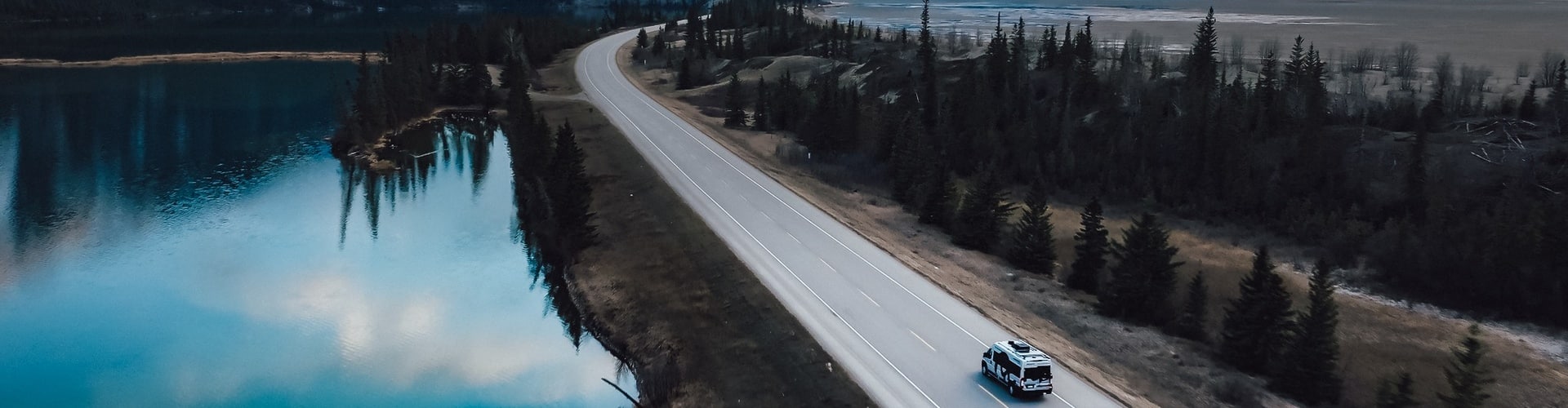 Aerial view of an RV driving along a mountain highway beside a reflective blue lake, illustrating the freedom of exploring different RV classes.