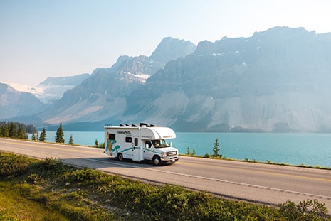 Scenic mountain landscape with a white roadsurfer RV driving along a highway beside a turquoise lake, surrounded by lush greenery and towering peaks with patches of snow