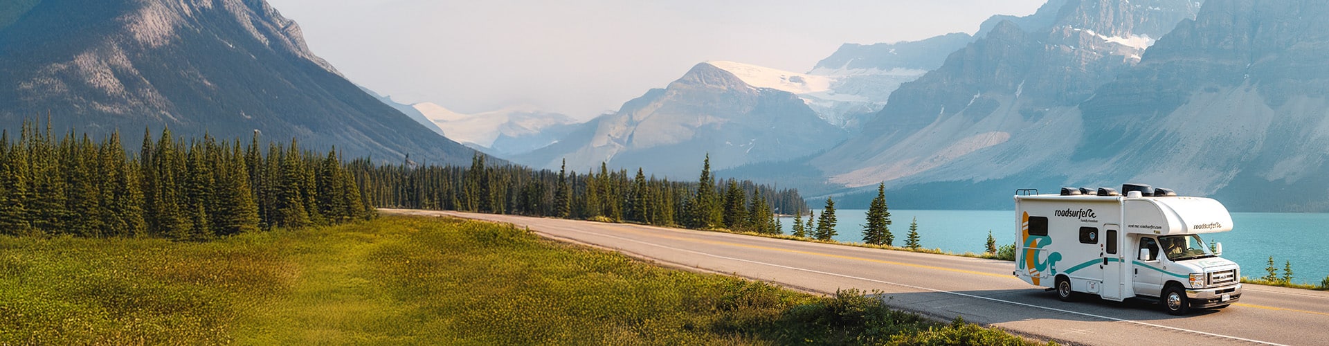 Scenic mountain landscape with a RV driving along a highway beside a turquoise lake, surrounded by lush greenery and towering peaks with patches of snow