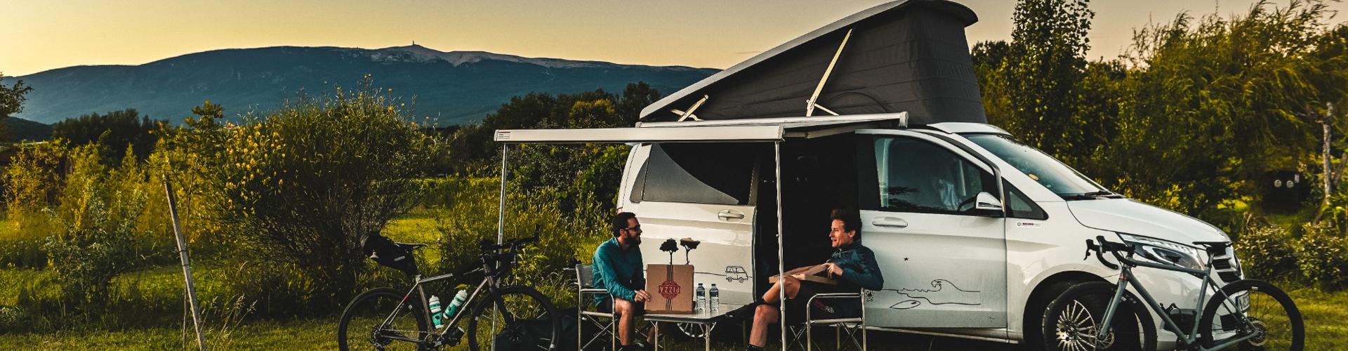 Two travelers relaxing beside their camper at an RV park during sunset, with open fields and distant mountains in the background.