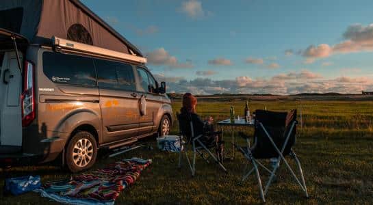 A peaceful RV park scene at sunset with a camper, camping chairs, and open fields in the background.