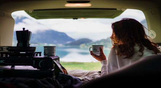 Woman relaxing in the back of a camper with a coffee mug, looking out over a lake and mountains, showing RV rentals near me for peaceful getaways