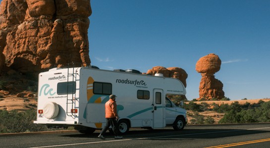 Class C RV parked on a desert highway beside red rock formations, showing RV rentals near me on a road trip