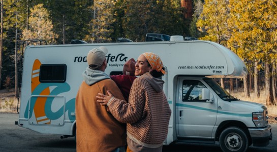 Family standing in front of a Class C RV with more of the vehicle and road visible, representing RV rentals near me for forest getaways