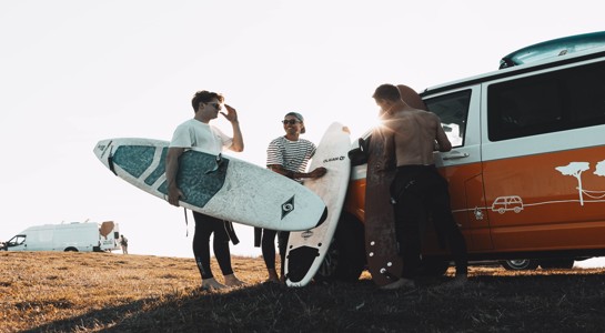 Group of friends with surfboards standing beside an orange camper van at the coast, showing RV rentals near me for beach trips